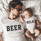 Man wearing a 'BEER' shirt and holding a baby in a 'MILK' onesie on a bed.