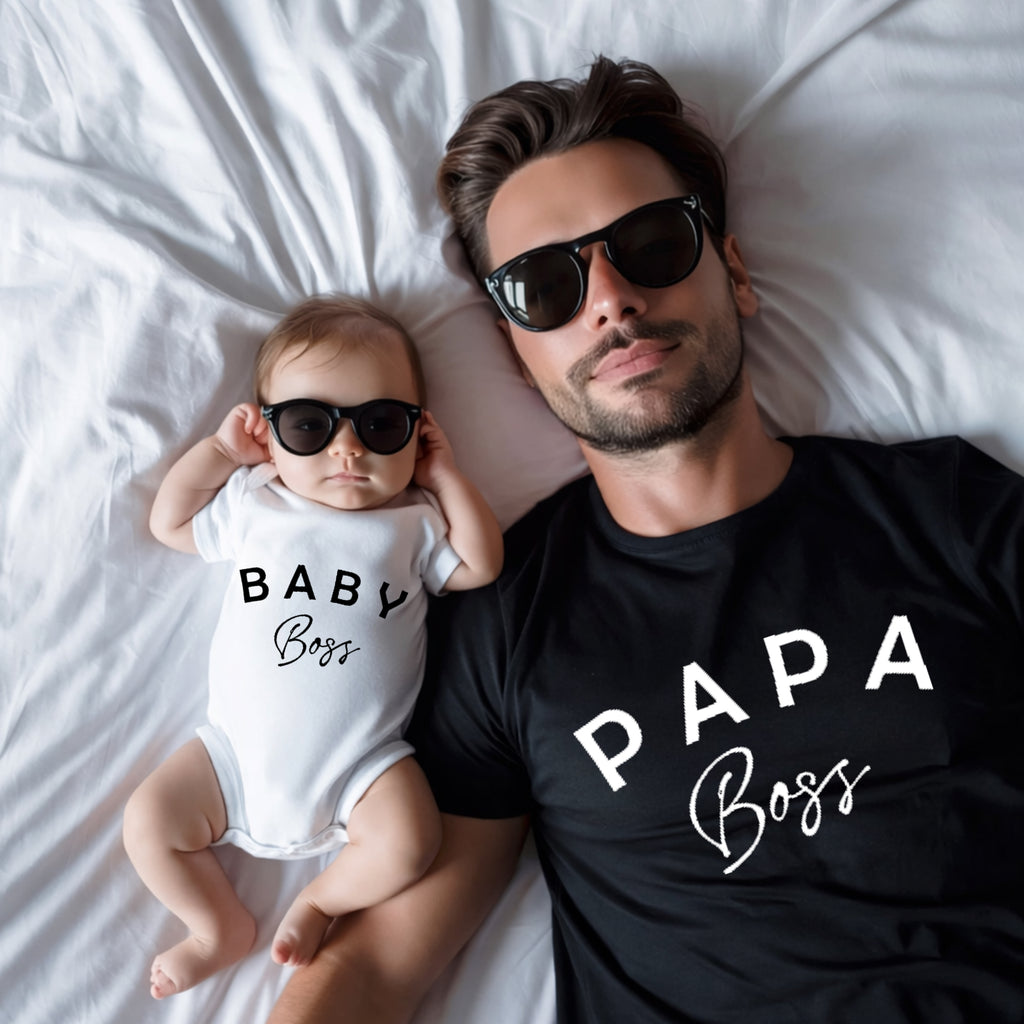 Man and baby wearing sunglasses and 'Papa Boss' and 'Baby Boss' shirts on a bed.