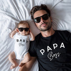 Man and baby wearing sunglasses and 'Papa Boss' and 'Baby Boss' shirts on a bed.