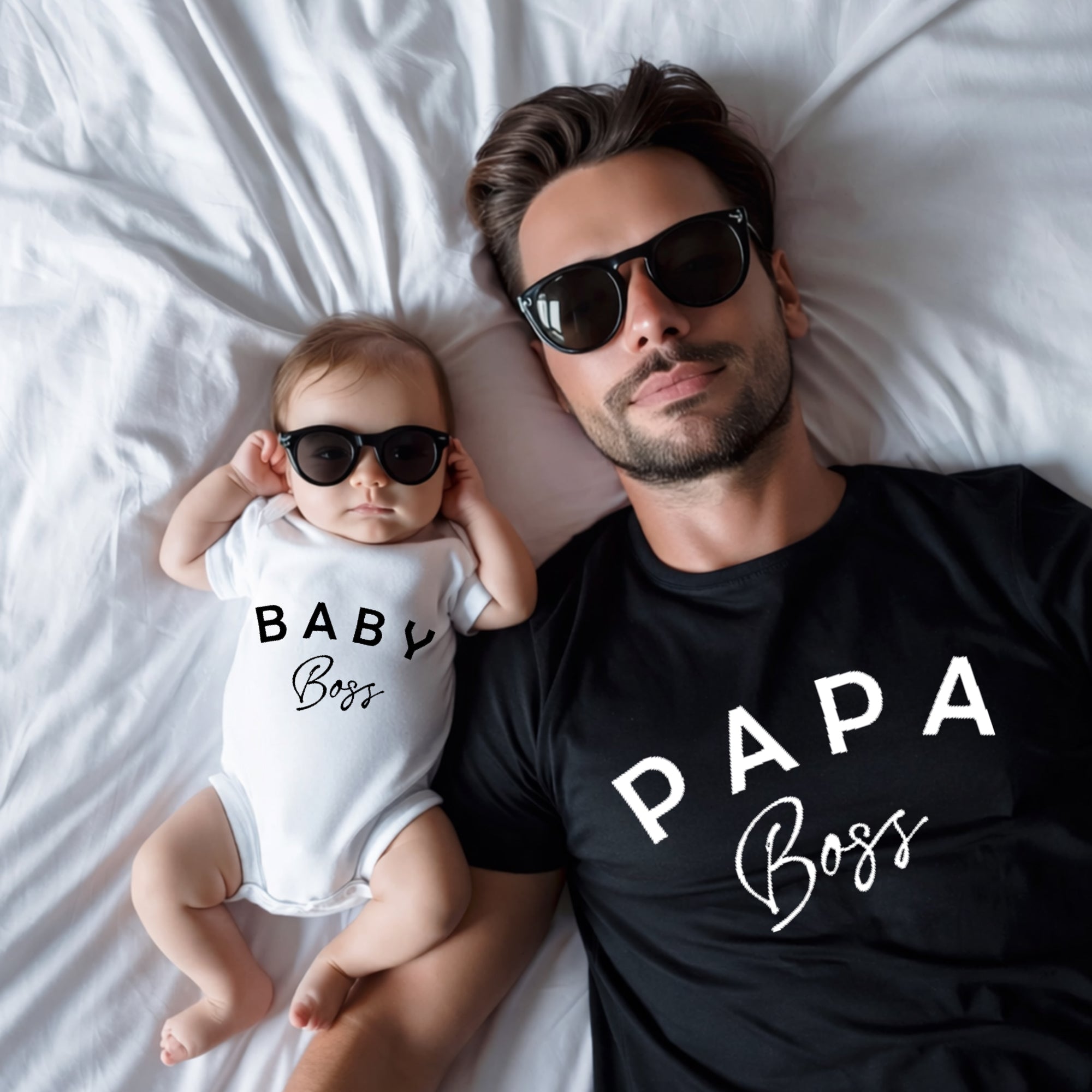 Man and baby wearing sunglasses and 'Papa Boss' and 'Baby Boss' shirts on a bed.