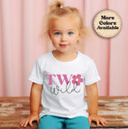Child wearing a white t-shirt with 'TW wild' design on a wooden surface with pink curtain background